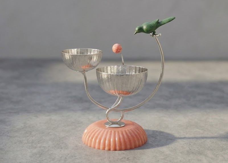 A decorative two-bowl metal candle holder with a ribbed peach-pink base, small peach-colored beads inside the bowls, a green bird figurine perched on the right arm, and a single bead suspended above the center, set against a neutral gray background.
