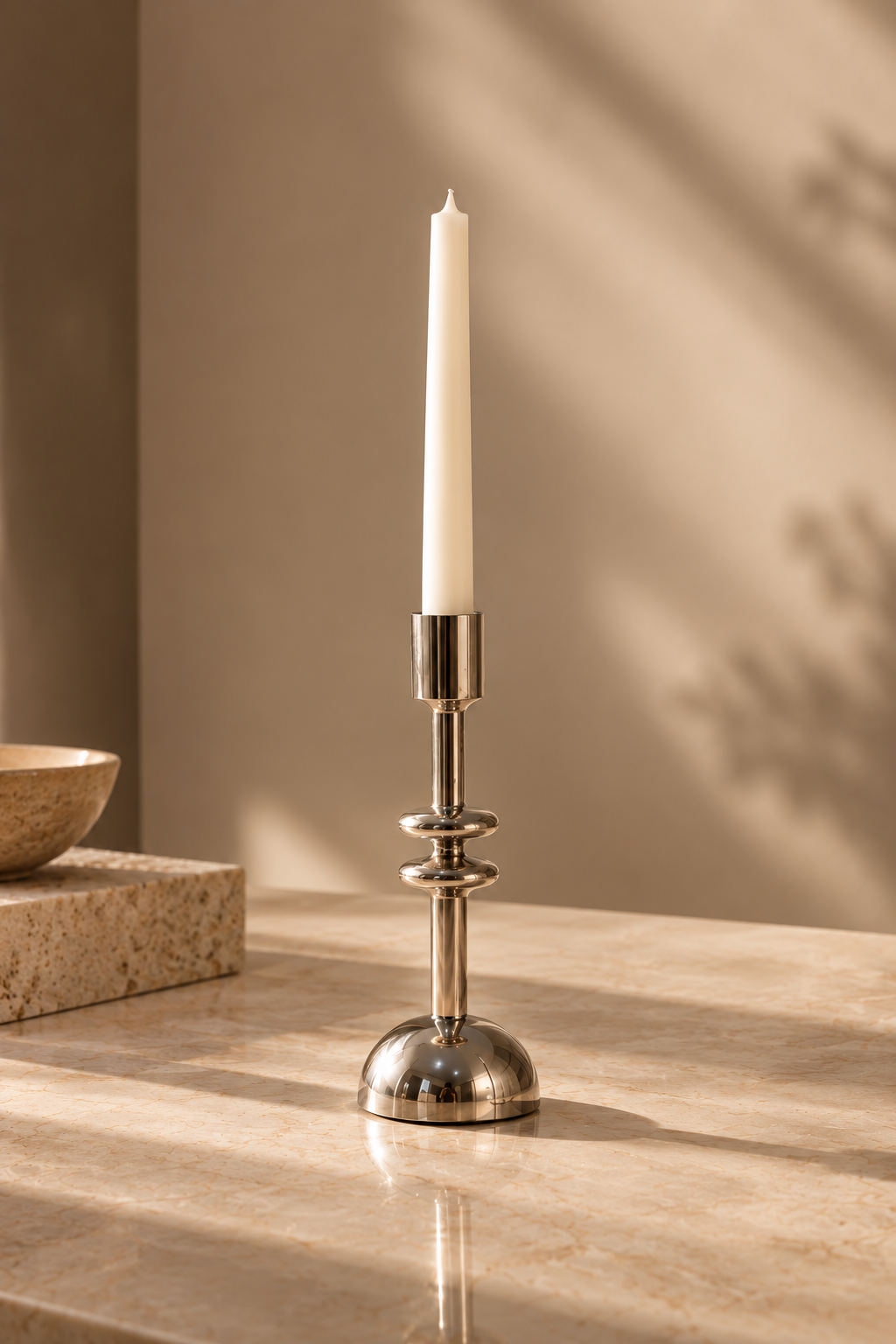 A polished silver metal candlestick holder with a white taper candle, set on a beige marble surface with warm sunlight and soft shadows.