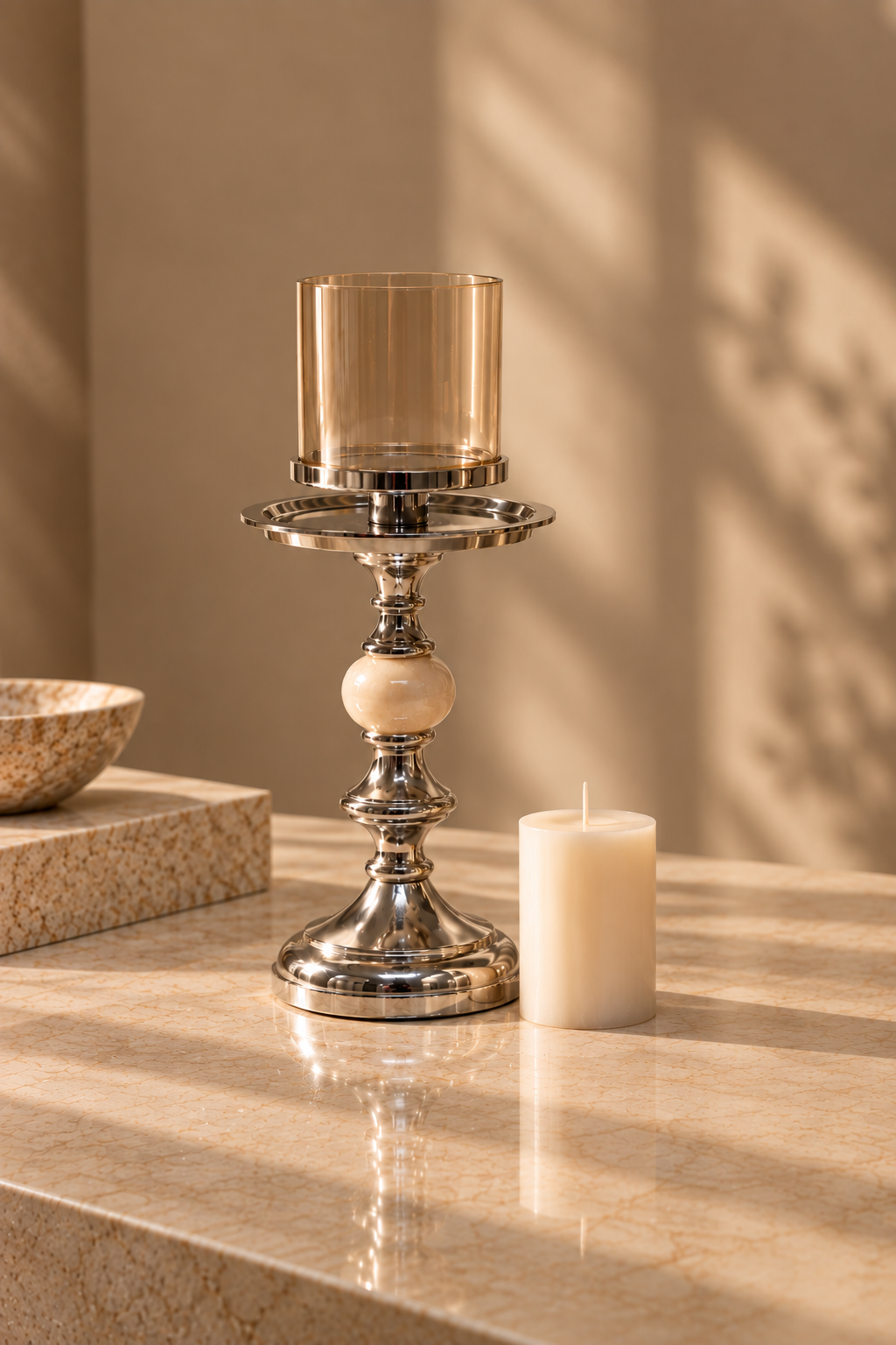 A polished silver-toned metal candlestick holder with a clear amber glass candle holder and a white pillar candle, styled on a beige marble surface with warm sunlight and leaf shadows.