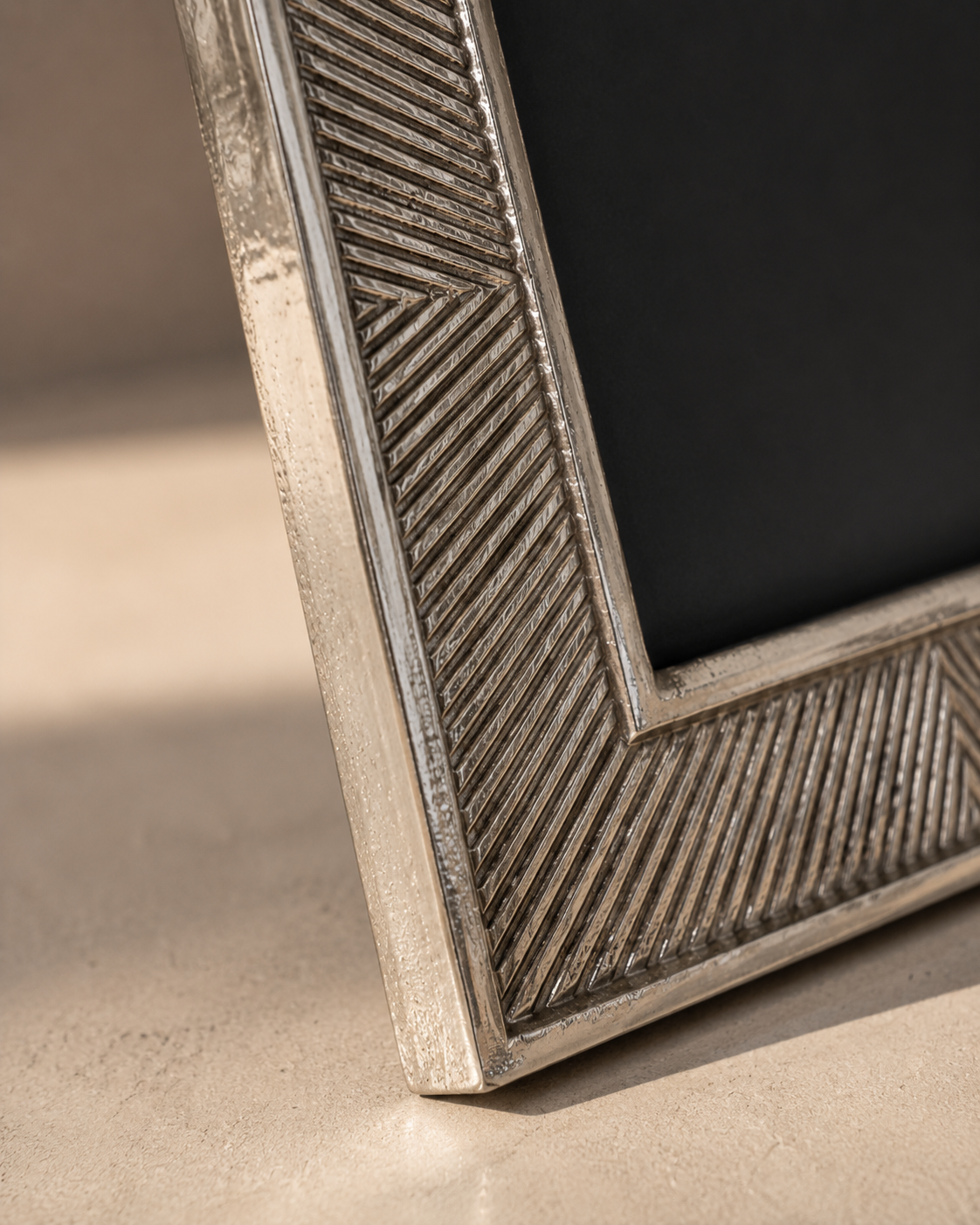 Close-up of a textured silver picture frame corner with a geometric herringbone pattern, resting on a beige surface with a dark background.