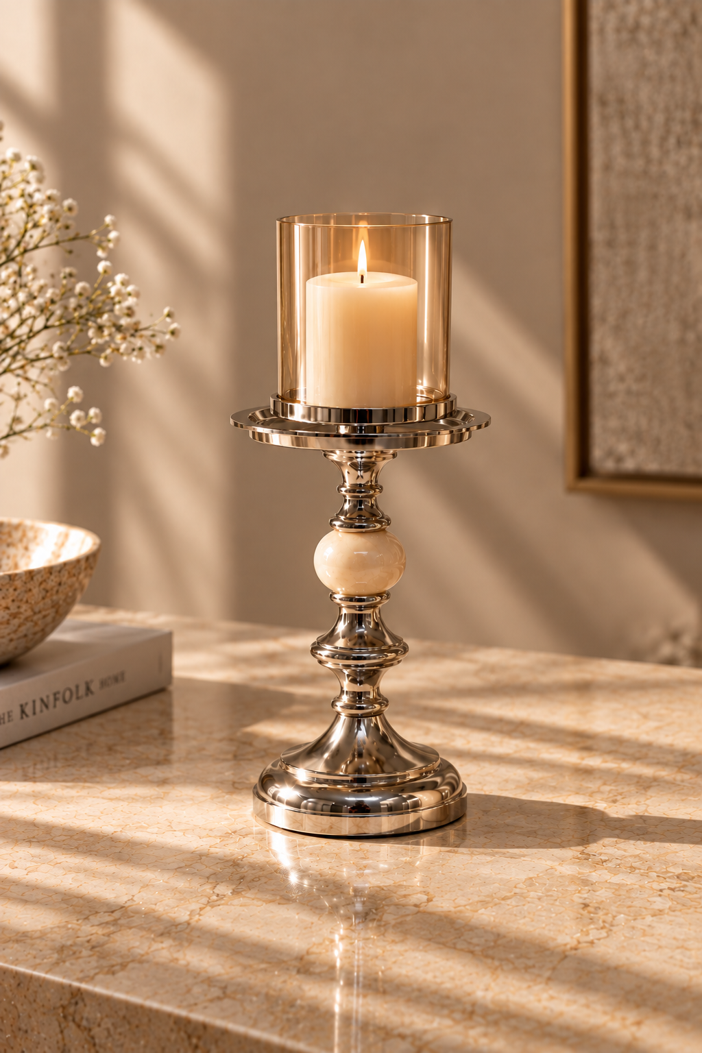 A silver-toned metal candlestick holder with a clear glass cylinder and lit cream candle, placed on a beige stone surface in a warm interior setting with sunlight and shadows.