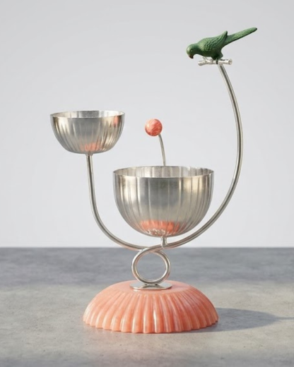 A decorative metal candle holder with two silver bowls on curved supports, a small pink spherical accent, and a green bird figurine perched at the top, set on a ribbed peach-pink base against a light gray background.