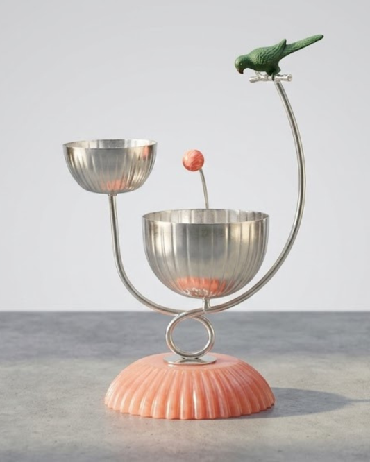 A decorative metal candle holder with two silver bowls on curved supports, a small pink spherical accent, and a green bird figurine perched at the top, set on a ribbed peach-pink base against a light gray background.