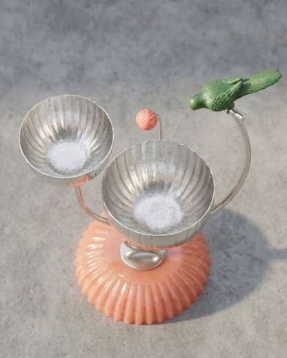 A decorative metal candle holder with two ribbed silver bowls on curved stems, mounted on a scalloped peach-pink base and topped with a green bird figurine, set against a textured gray background.