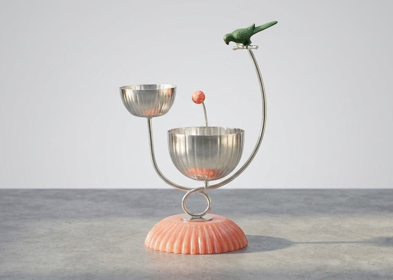 A decorative metal and wood bird feeder with a ribbed pink base, two silver bowls, a curved silver stand, and a small green bird perched on the top right.