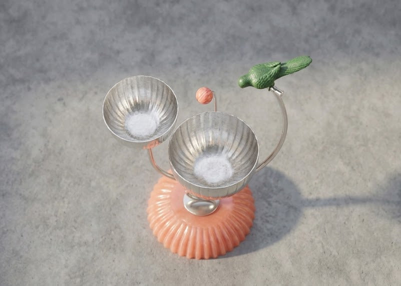 A decorative tabletop sculpture with two ribbed metallic bowls on curved stems, a small green bird perched on one stem, a pink orb, and a ribbed peach-pink base, shown on a gray textured surface.