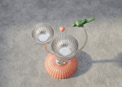 A decorative tabletop sculpture with two ribbed metallic bowls on curved stems, a small green bird perched on one stem, a pink orb, and a ribbed peach-pink base, shown on a gray textured surface.