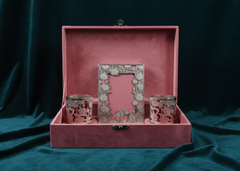 A pink velvet presentation box opened on a dark teal fabric backdrop, containing a floral-patterned picture frame and two matching pink patterned candle holders.