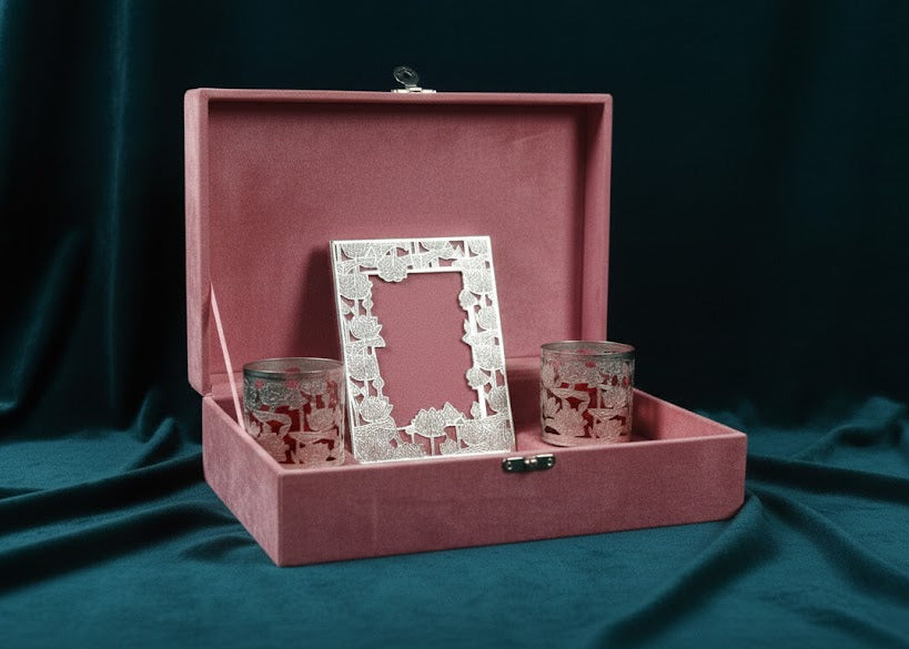 A pink velvet presentation box with a hinged lid, shown open on a dark teal fabric backdrop. Inside the box are a white floral-patterned picture frame and two matching pink-and-white patterned glass cups or candleholders.