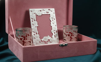 A pink velvet presentation box with a hinged lid, holding a silver floral-patterned picture frame and two matching pink-and-silver etched glass cups, set against a dark teal fabric background.