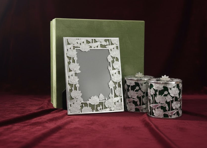A decorative silver picture frame with a floral cutout border stands in front of a green box, with two matching patterned candle holders beside it on a dark red fabric background.