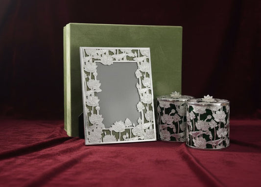 A decorative silver picture frame with a floral cutout border stands in front of a green box, with two matching patterned candle holders beside it on a dark red fabric background.