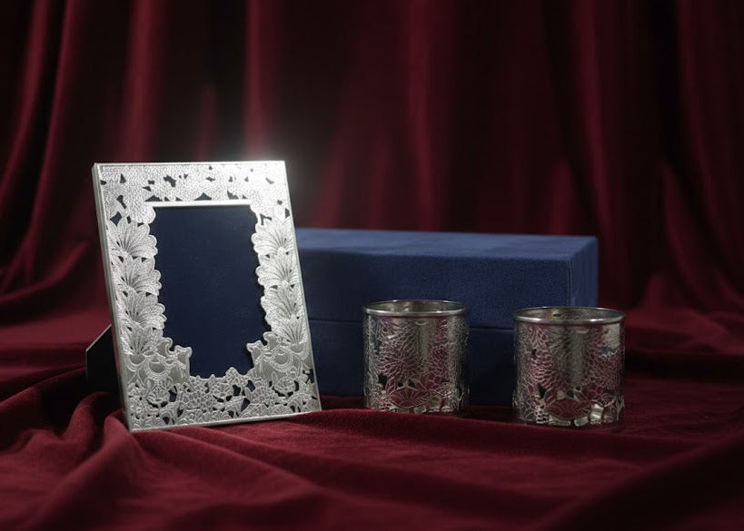 A silver floral-patterned picture frame with a dark insert, displayed next to two matching silver candle holders and a blue velvet box on draped red fabric.