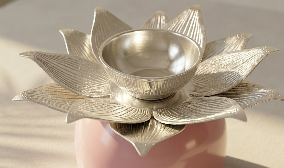 A silver-toned metal lotus-shaped holder with a small central bowl, displayed on a soft pink base against a light neutral background.