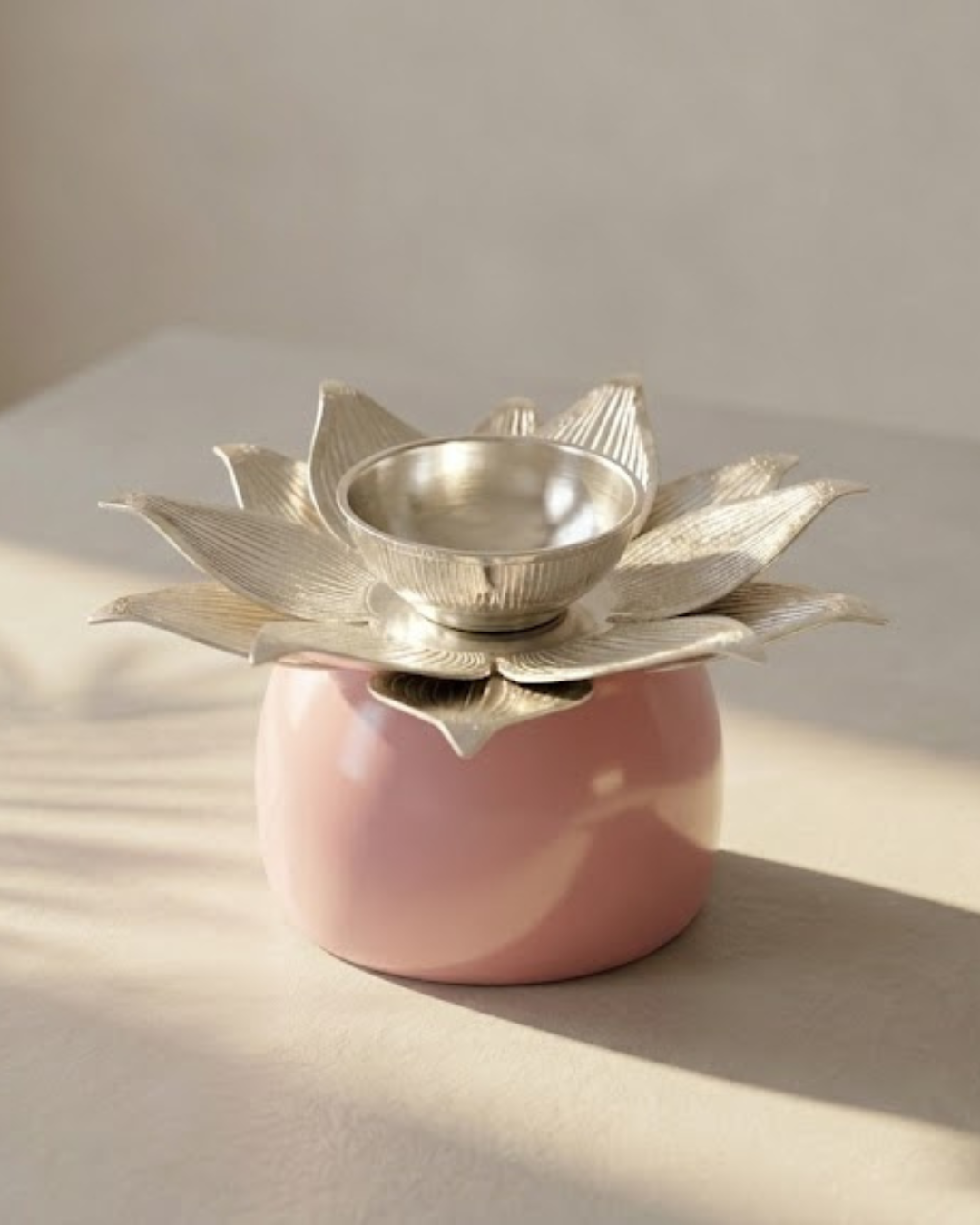 A pink ceramic oil lamp holder with a lotus-shaped silver metal top and a small silver bowl, photographed on a warm beige surface with soft sunlight and shadows.