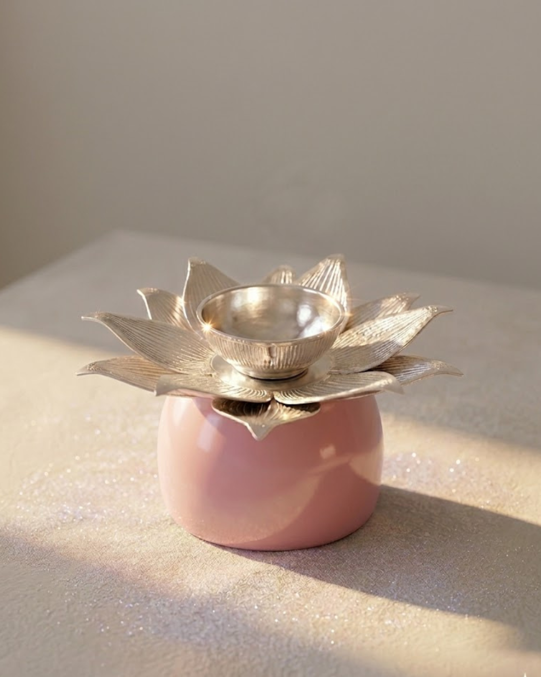 A pink ceramic oil lamp holder with a silver-toned lotus flower top and a small metallic bowl, photographed on a neutral beige surface with soft natural light.