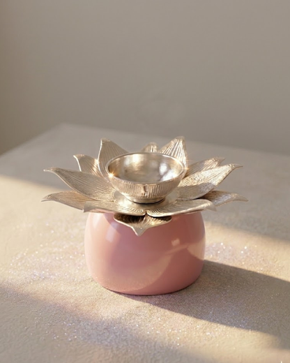 A pink ceramic oil lamp holder with a silver-toned lotus flower top and a small metallic bowl, photographed on a neutral beige surface with soft natural light.