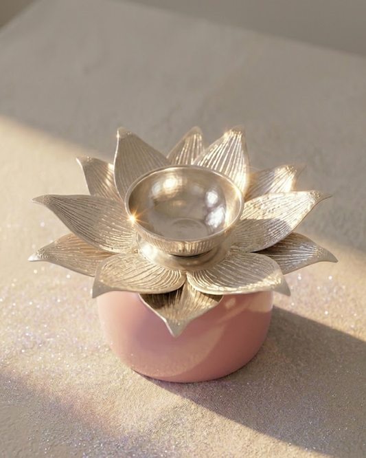 A metallic silver lotus-shaped candle holder with a small reflective bowl in the center, set on a round pink base against a glittery beige background.