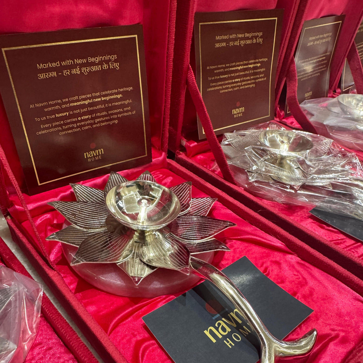 A silver-toned decorative diya holder with a lotus-shaped bowl and curved handle, presented in a red velvet box with an insert card and black brand tag. Multiple matching boxes are visible in the background.
