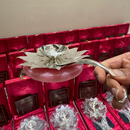 A hand holding a silver-toned lotus-shaped candle holder over a pink glass dish, with multiple matching silver lotus candle holders displayed in red presentation boxes in the background.