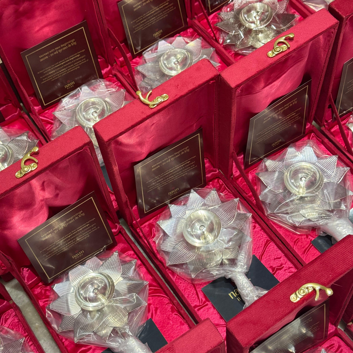 Multiple silver-toned decorative hand fans with clear plastic wrapping and gold-tone clasps, arranged inside red velvet gift boxes with black plaques.