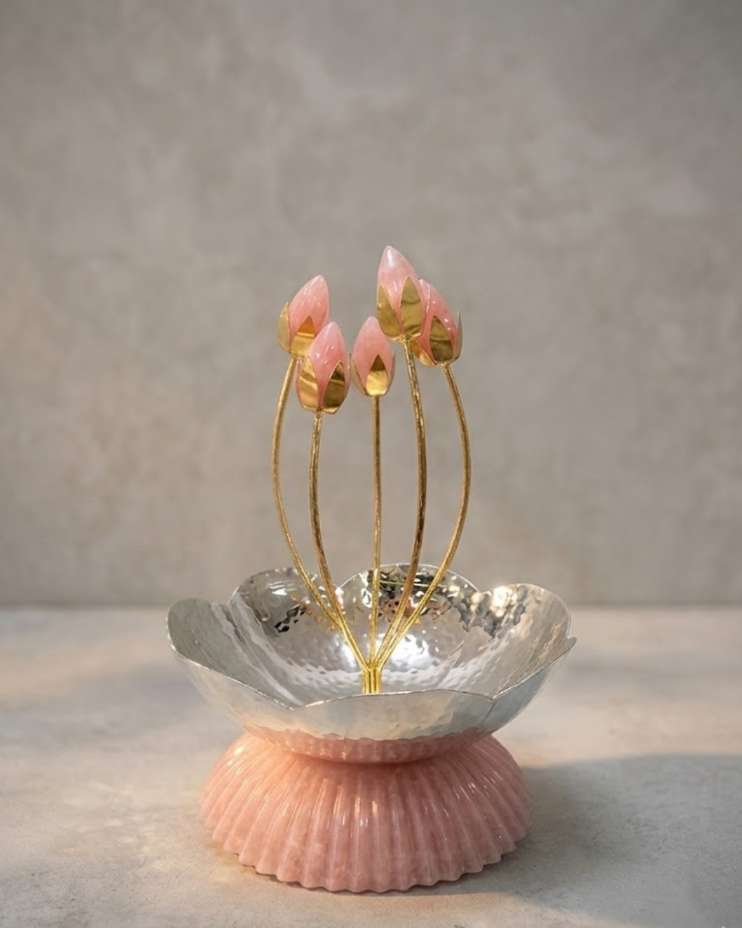 A decorative lotus flower holder featuring five pink lotus buds on gold-colored stems, set in a hammered silver metal cup atop a ribbed pink base, photographed against a neutral textured gray background.