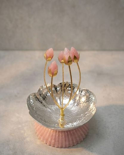 A decorative jewelry stand featuring pink rosebud-shaped earrings on gold-tone stems, set in a hammered silver-tone bowl with a pink ribbed base, photographed on a neutral gray surface.