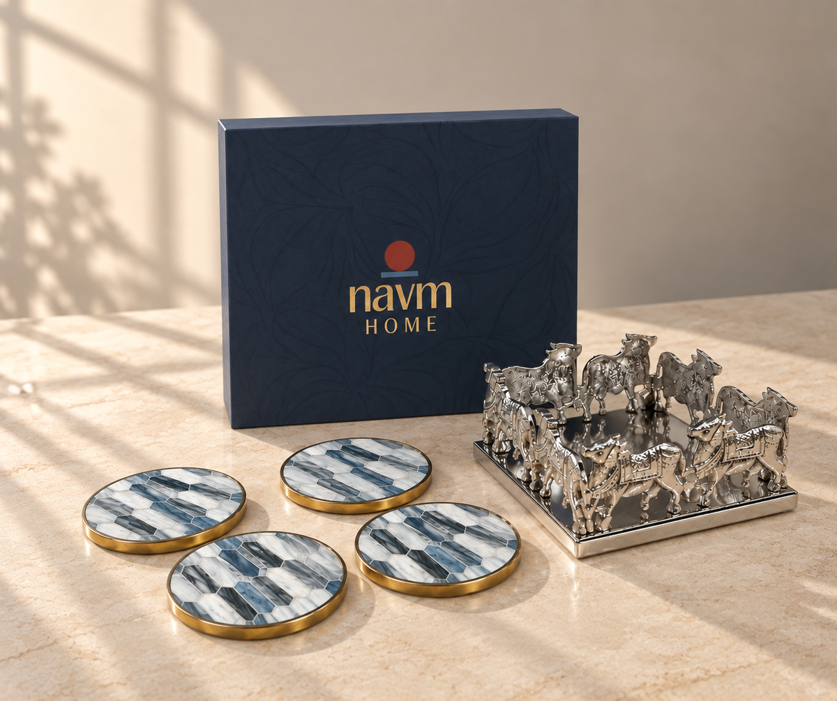 Navm Home gift set featuring a dark navy blue box, four round coasters with blue and white marble patterns and gold trim, and a silver horse figurine tray, arranged on a beige marble surface with warm natural light shadows.