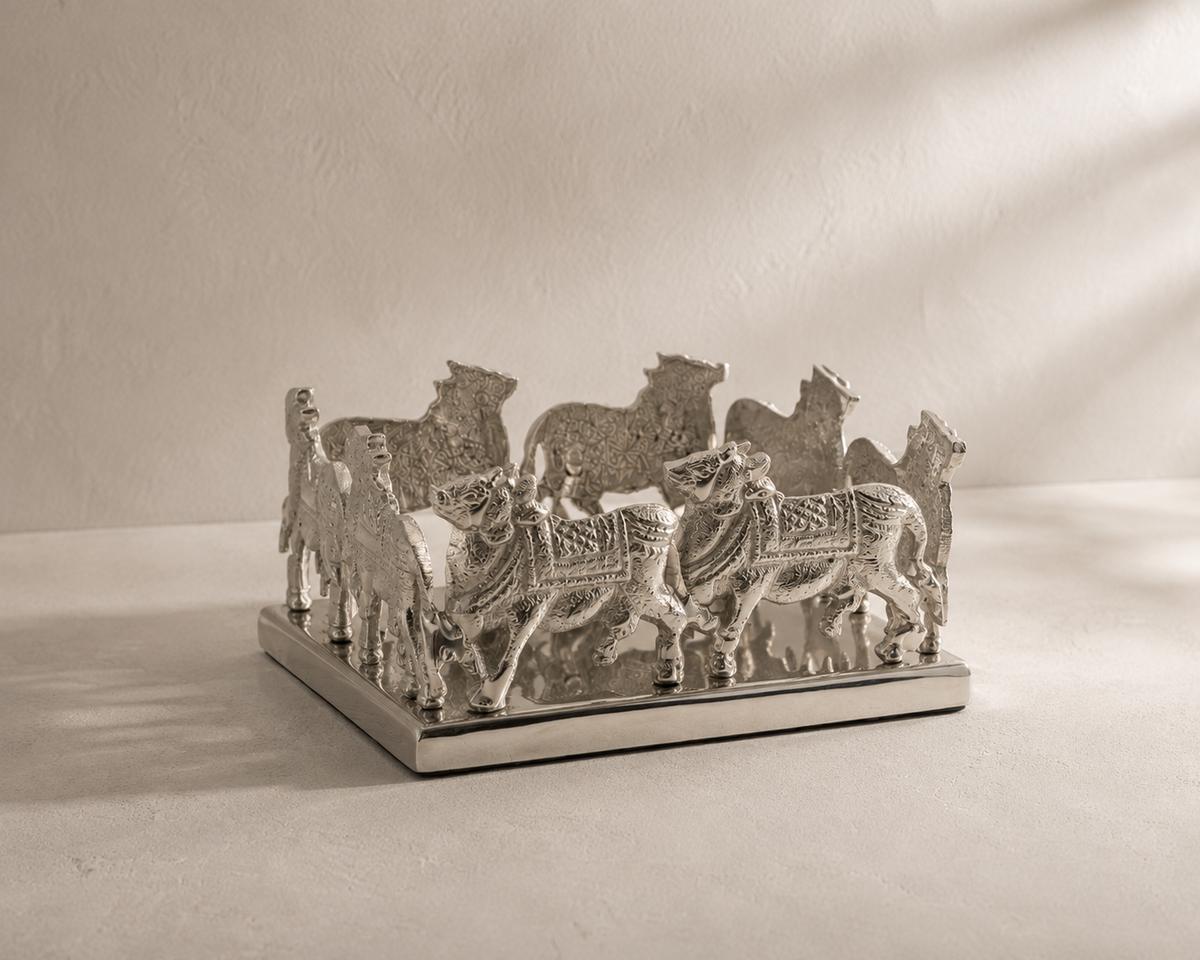A silver-toned decorative sculpture featuring multiple stylized animal figures, of nandi cow, standing on a rectangular base against a textured beige wall and floor with dramatic diagonal shadows.
