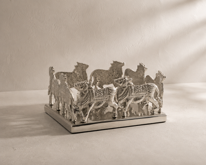 A silver-toned decorative sculpture featuring multiple stylized animal figures, of nandi cow, standing on a rectangular base against a textured beige wall and floor with dramatic diagonal shadows.