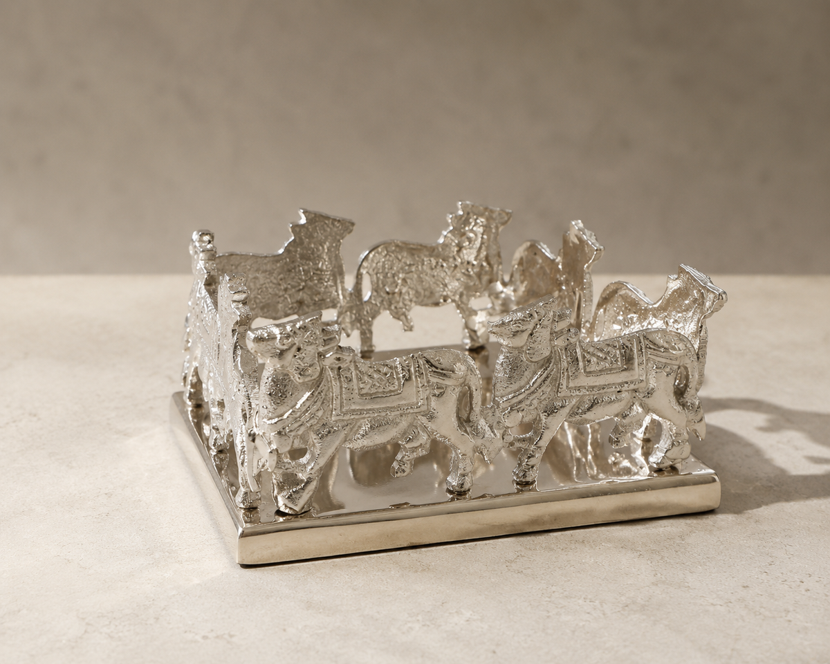 A polished silver decorative sculpture featuring a herd of cows standing on a rectangular base, photographed on a light stone surface against a neutral beige background.
