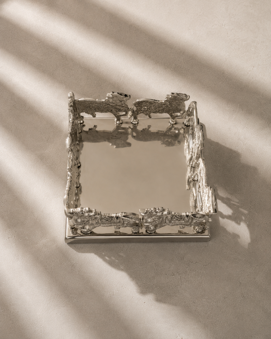 A rectangular mirrored tray with ornate, textured silver metal borders featuring sculpted animal figures, photographed on a neutral beige surface with dramatic diagonal shadows.