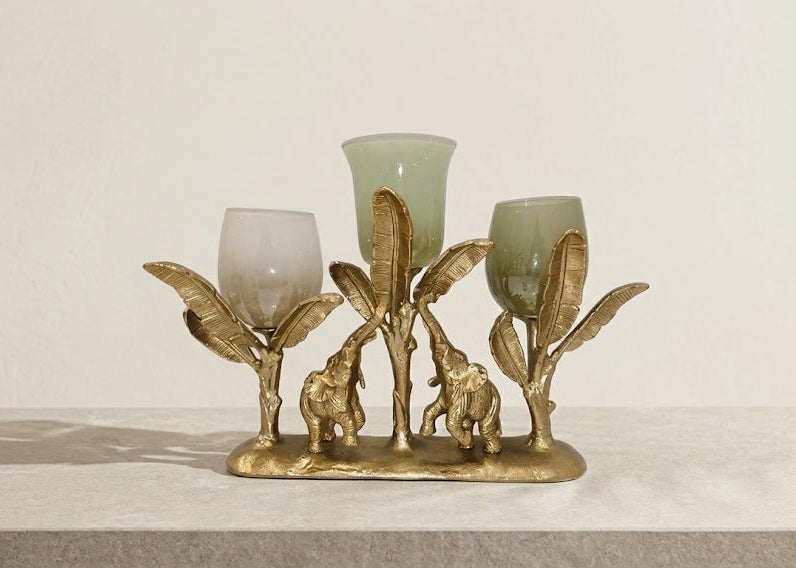 A decorative gold-toned metal candle holder featuring elephant and palm tree motifs, holding three translucent pale green glass candle cups, displayed on a light stone surface against a plain off-white wall.