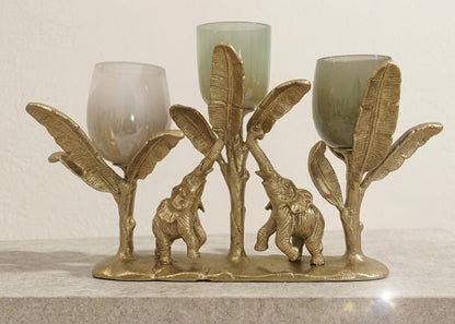 A decorative brass candle holder featuring elephant and banana leaf motifs, holding three glass candleholders, displayed on a stone shelf against a neutral wall.