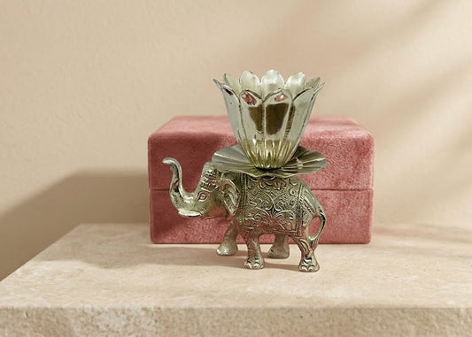 A small silver elephant figurine holding a lotus-shaped flower on its back, displayed in front of a pink velvet box on a beige stone surface against a beige wall.