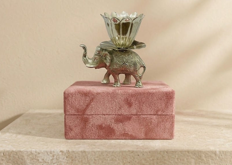 A small silver elephant figurine holding a floral cup, displayed on top of a pink velvet box against a warm beige background.