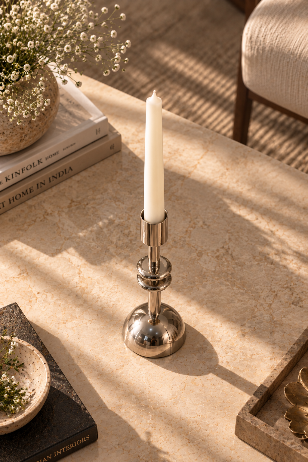A polished silver metal candlestick holder with a white taper candle, styled on a beige stone tabletop with warm sunlight and shadows.