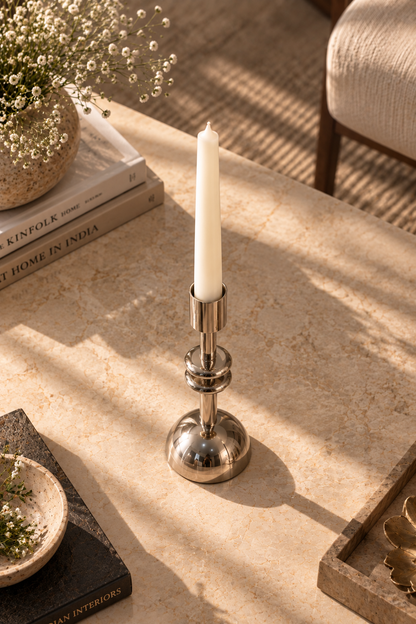 A polished silver metal candlestick holder with a white taper candle, styled on a beige stone tabletop with warm sunlight and shadows.