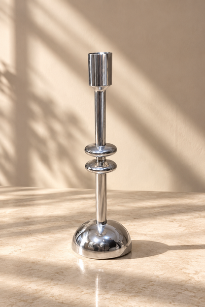 A polished silver metal candlestick holder with a flared round base and a cylindrical top, standing on a beige stone surface with warm sunlight and leaf shadows cast across the background.