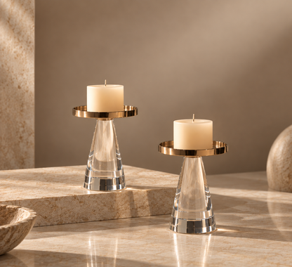 Two clear glass candle holders with gold-toned circular rings and cream pillar candles, styled on a polished stone surface against a warm beige background.