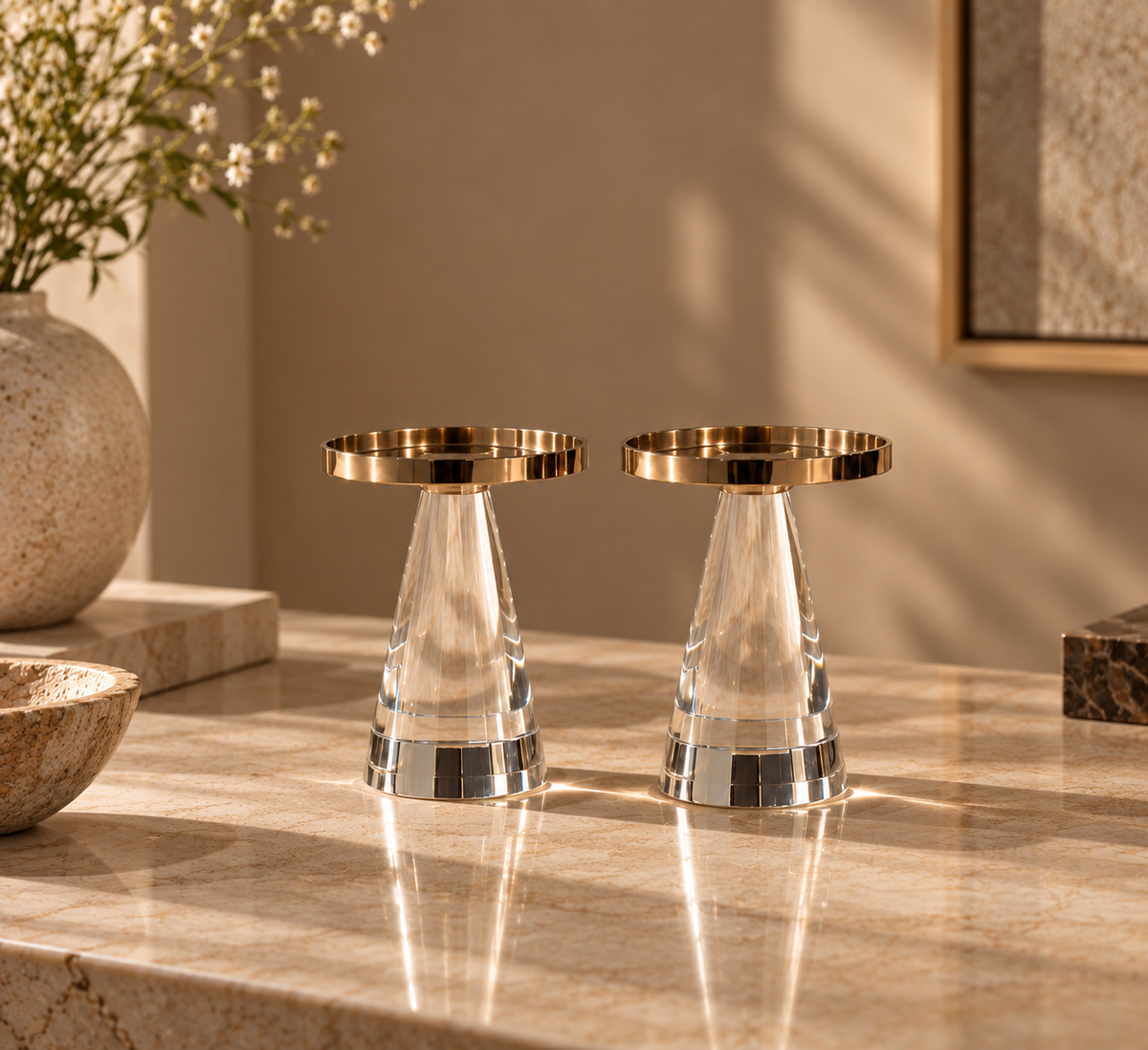 Two clear faceted crystal candle holders with gold-toned circular rims, displayed on a warm beige marble countertop in a styled interior setting.