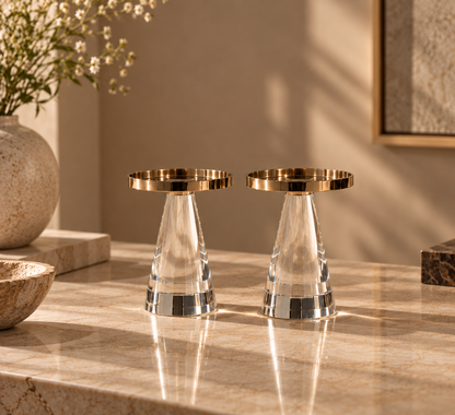 Two clear faceted crystal candle holders with gold-toned circular rims, displayed on a warm beige marble countertop in a styled interior setting.