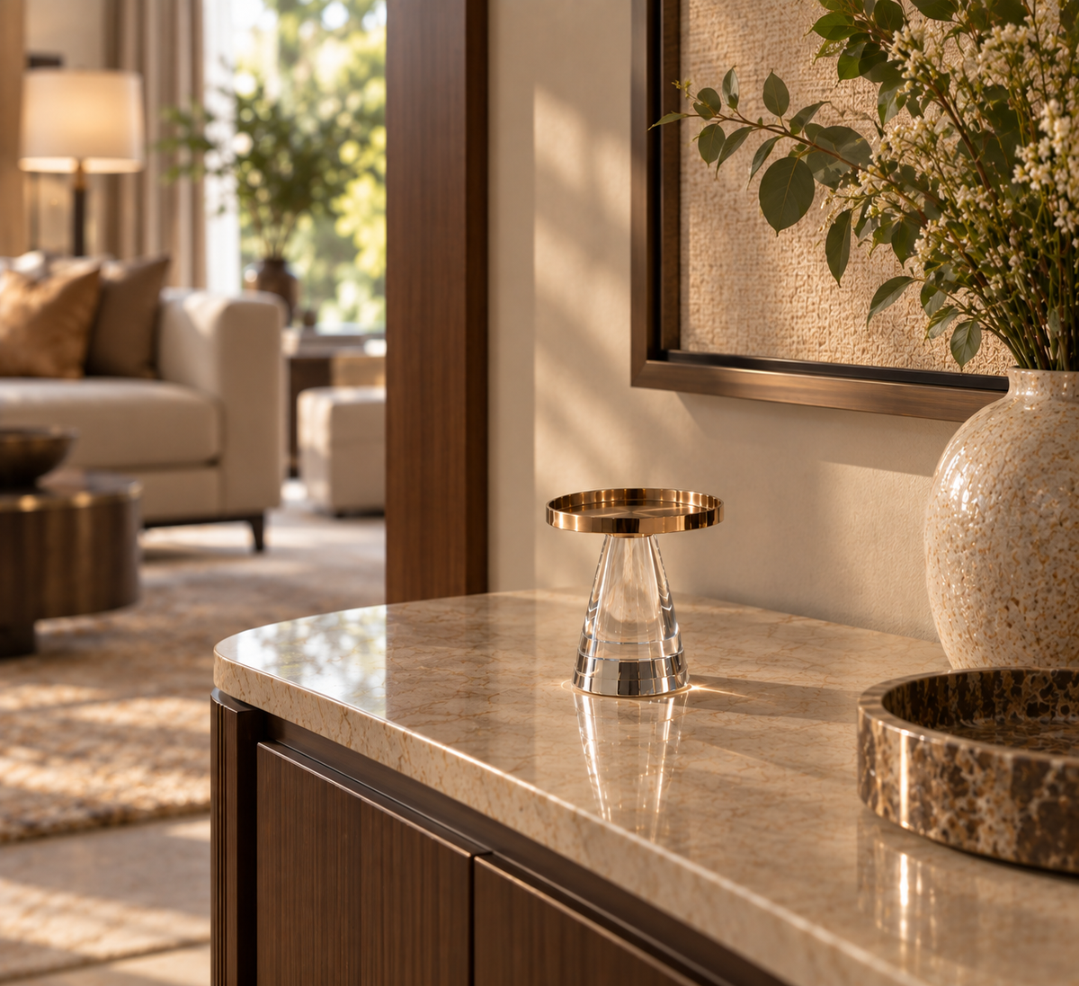 A small clear acrylic jewelry stand with a brushed gold circular top, displayed on a polished beige marble countertop in a modern, sunlit living room setting.