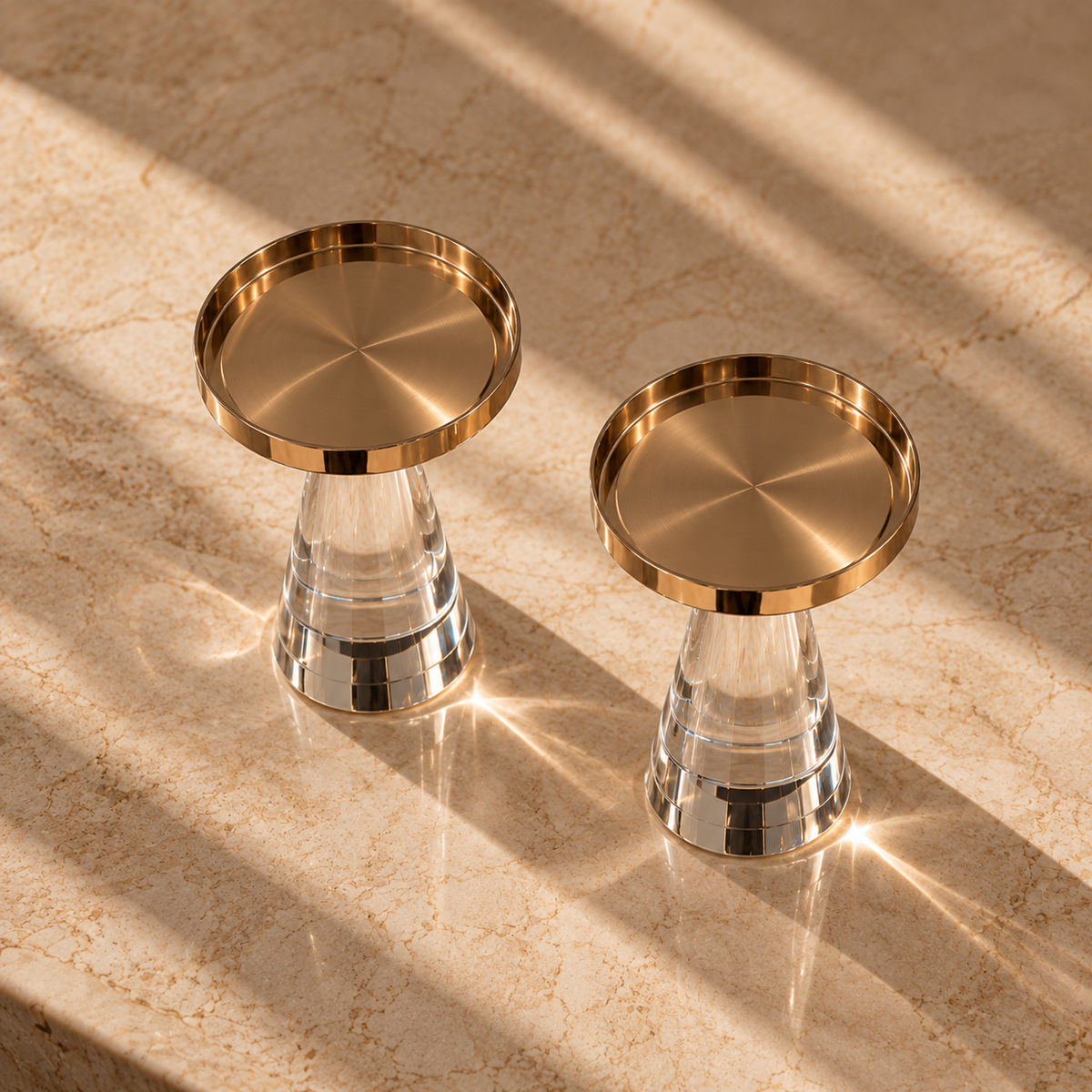 Two modern crystal candle holders with brushed gold metal rims and clear glass bases, styled on a beige marble surface with dramatic diagonal sunlight and shadows.