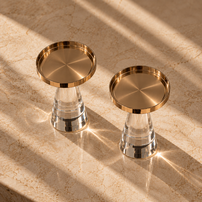Two modern crystal candle holders with brushed gold metal rims and clear glass bases, styled on a beige marble surface with dramatic diagonal sunlight and shadows.