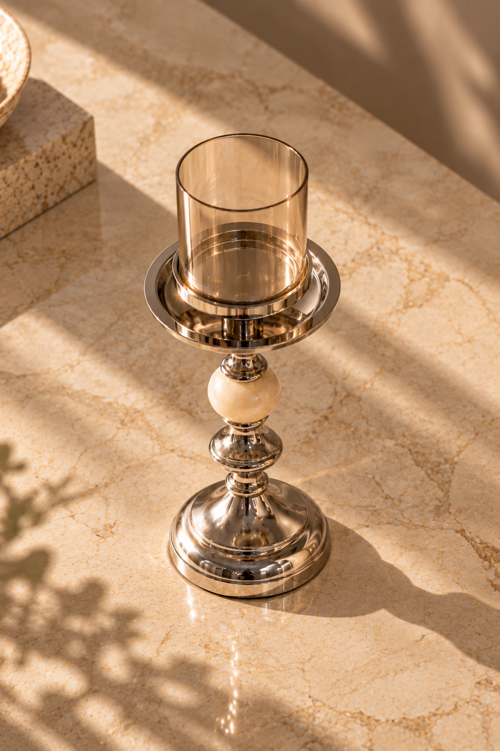 A polished silver-toned candlestick holder with a clear smoky amber glass candle holder, set on a beige marble surface with warm sunlight and leaf shadows.