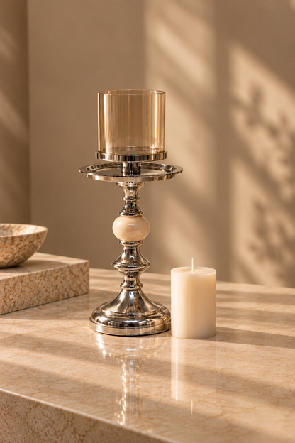 A polished silver-toned metal candlestick holder with a clear amber glass candle holder and a white pillar candle, styled on a beige marble surface with warm sunlight and leaf shadows.