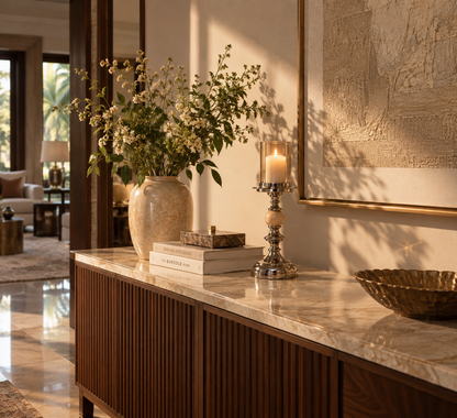 A warm, elegant interior scene featuring a wooden console table with a marble countertop, styled with a large ceramic vase of white flowers, books, a lit candle in a silver holder, and a gold bowl. Sunlight casts soft shadows across the wall and floor.