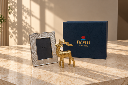 A gold-toned deer figurine and a silver textured picture frame displayed on a beige marble surface next to a navy blue 'navm HOME' gift box in a sunlit interior setting.