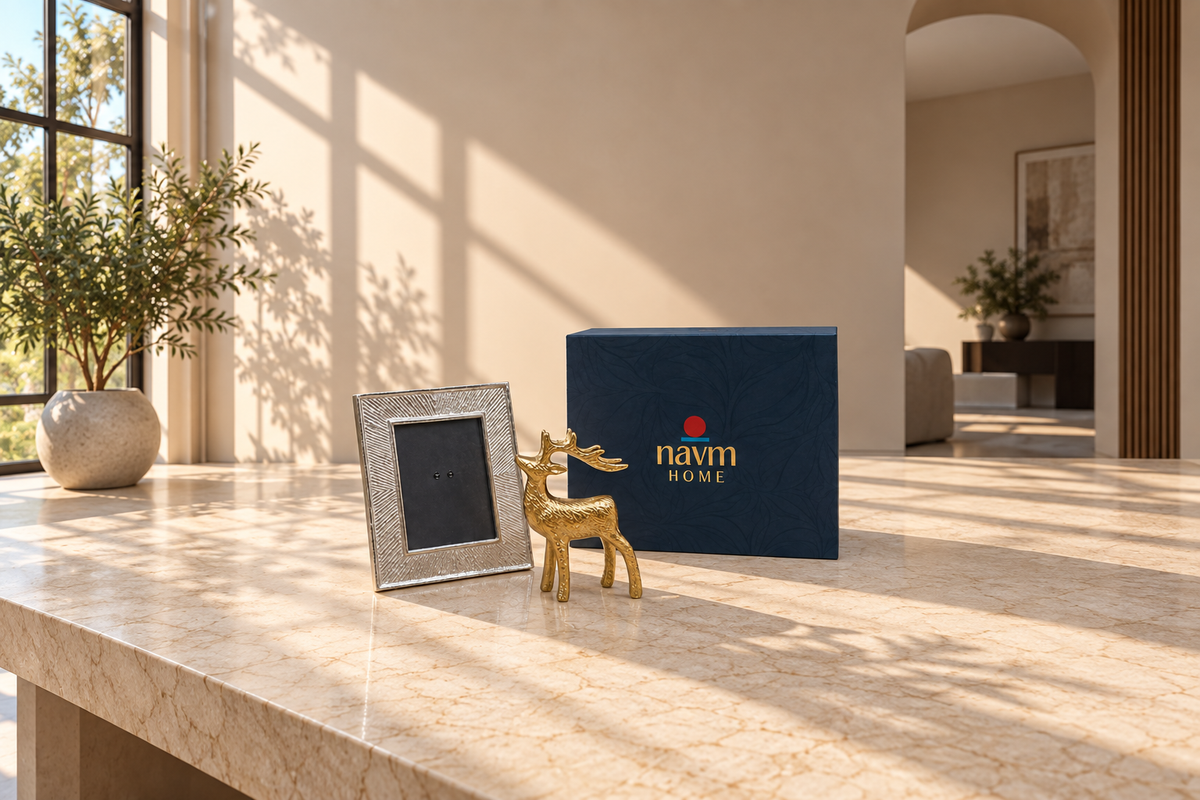 A navy blue gift box with the brand name 'navm HOME' in gold lettering sits on a marble countertop next to a small gold deer figurine and a silver picture frame, set in a beautifully lit modern living room.